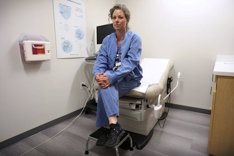 A woman wearing blue scrubs sits at the edge of a medical examining bed in an empty room.