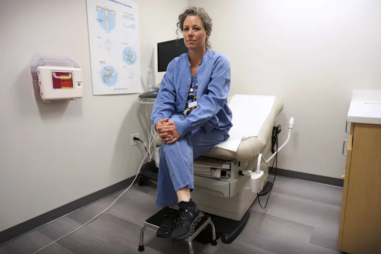 A woman wearing blue scrubs sits at the edge of a medical examining bed in an empty room.