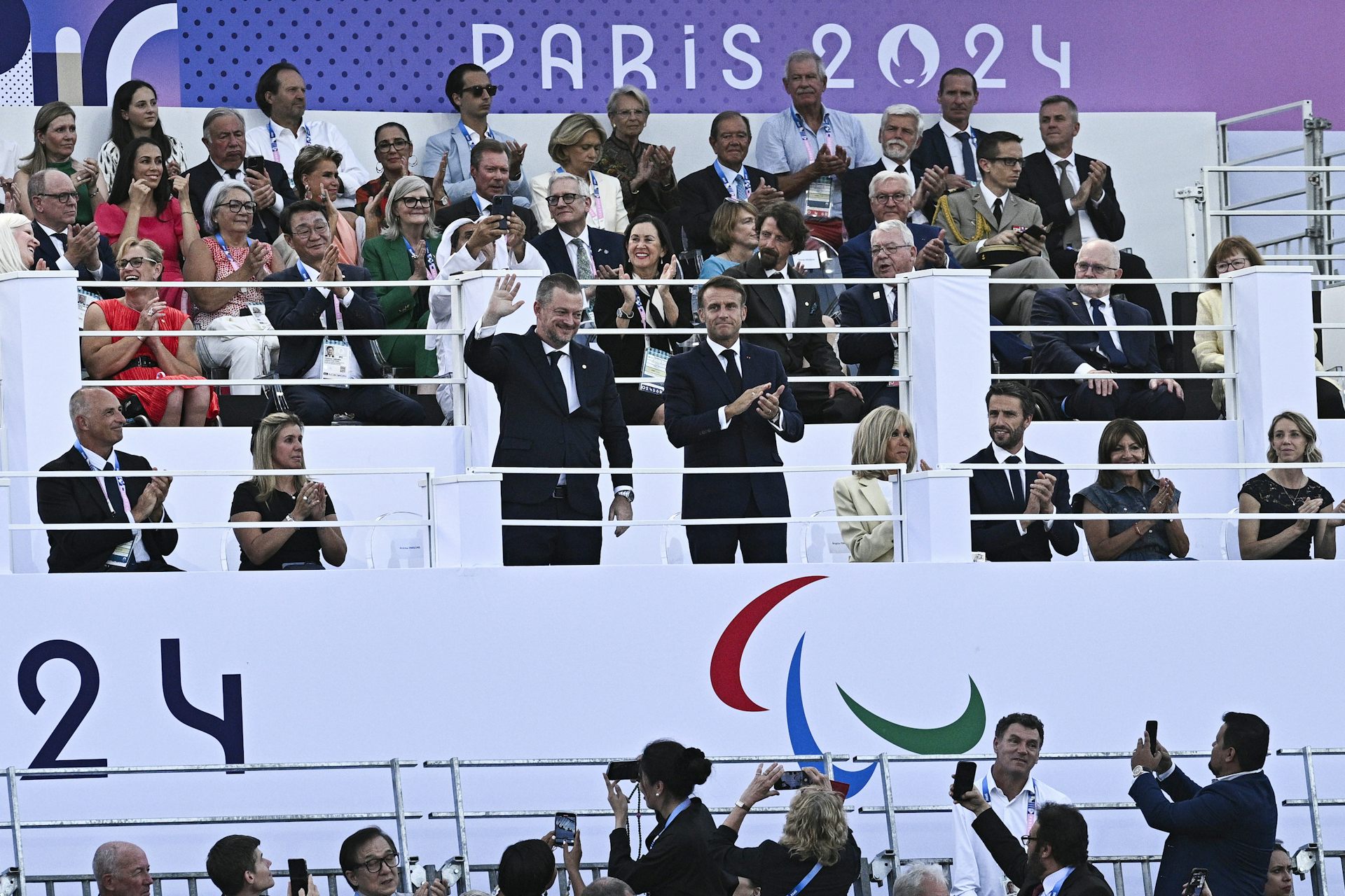 Two men in suits stand, one clapping while the other waves, in the stands of a stadium. Several other people in suits sit while applauding beside them.