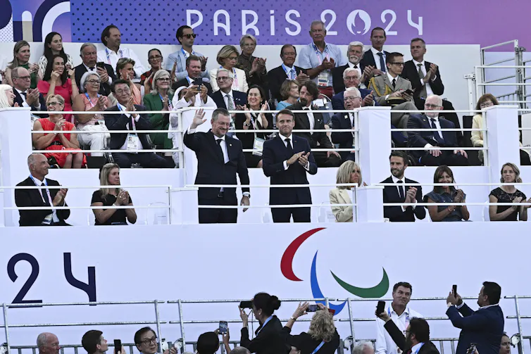 Two men in suits stand, one clapping while the other waves, in the stands of a stadium. Several other people in suits sit while applauding beside them.
