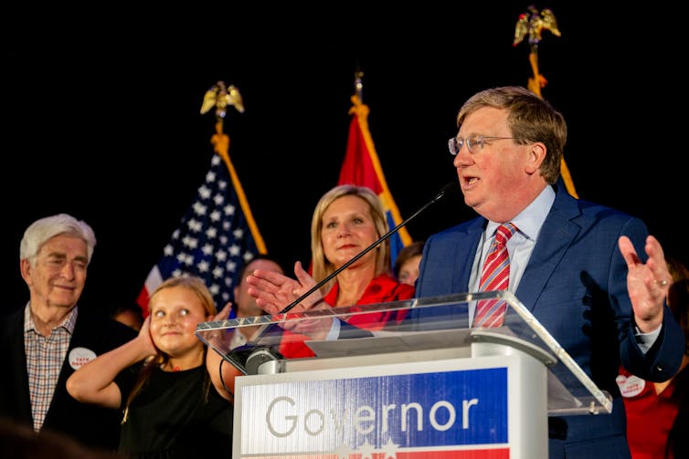 A middle-aged white man gestures to a crowd of supporters as he stands on a stage surrounded by family members.
