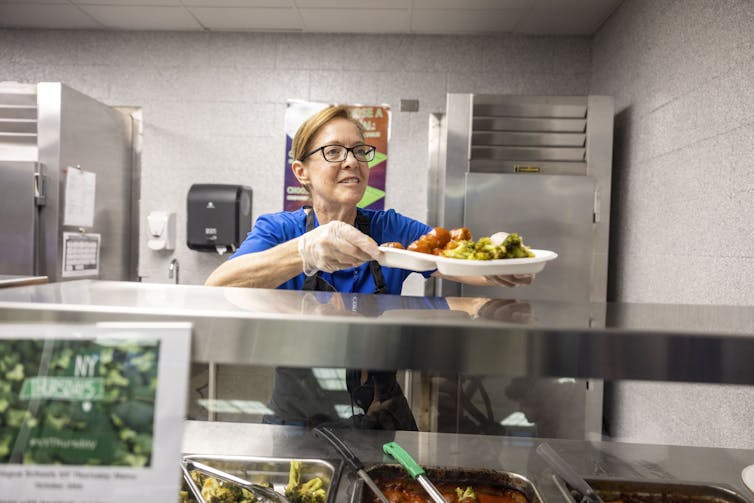 A woman in a cafeteria hands a heaping plate of food to the next person in line.