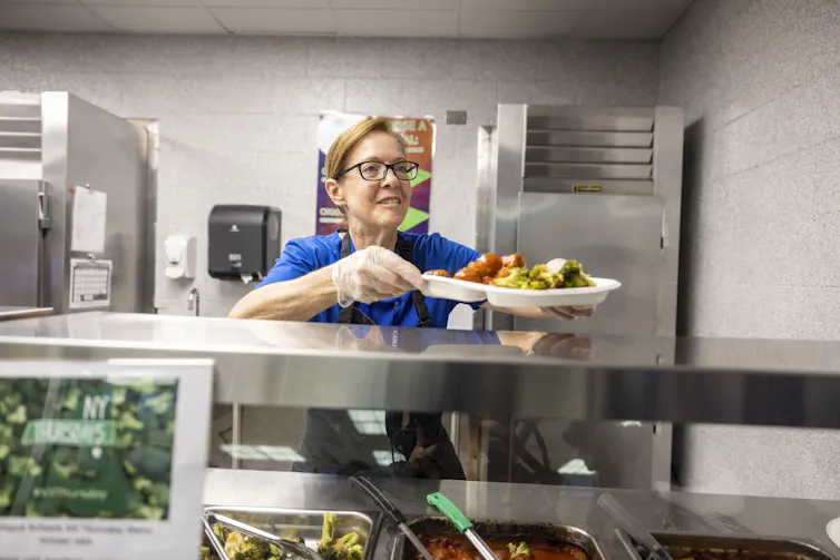 A woman in a cafeteria hands a heaping plate of food to the next person in line.