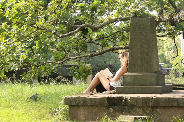 A woman reads a book as she sits next to a grave surrounded by trees