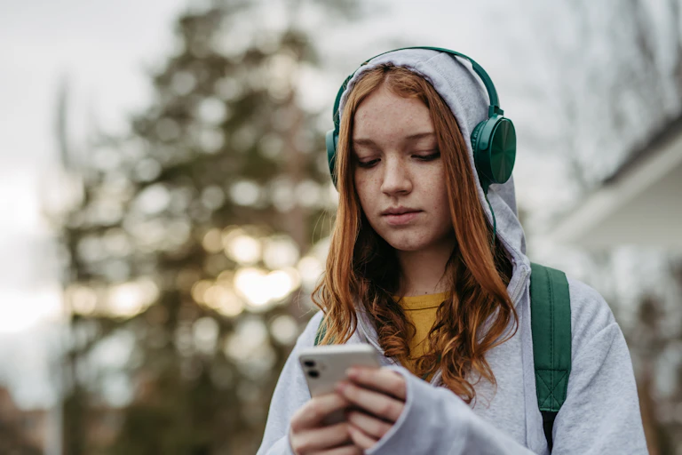 A teenage girls in headphones and hoodie holds a mobile phone.