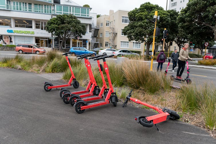 Shared e-scooters lined up on the edge of a road with people riding some behind them