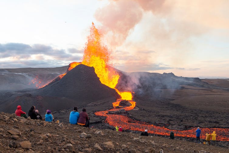 A small group of people sit on a rocky ridge watching a fiery volcano erupt in the distance with smoke and bright lava.