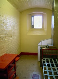 A small prison cell with white walls, a bed and table.