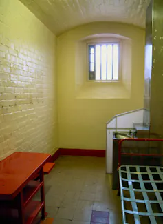 A small prison cell with white walls, a bed and table.