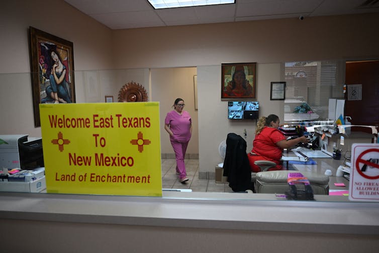 A yellow sign says 'Welcome East Texans to New Mexico' in front of two women wearing scrubs sitting at a desk and walking.