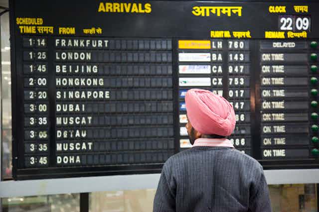 A man wearing a pink turban looks at an airport arrivals screen. A man wearing a pink turban looks at an airport arrivals screen.