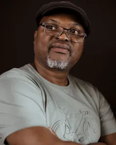 A portrait of an African man in glasses and a cap, his beard greying. He looks off camera with folded arms.