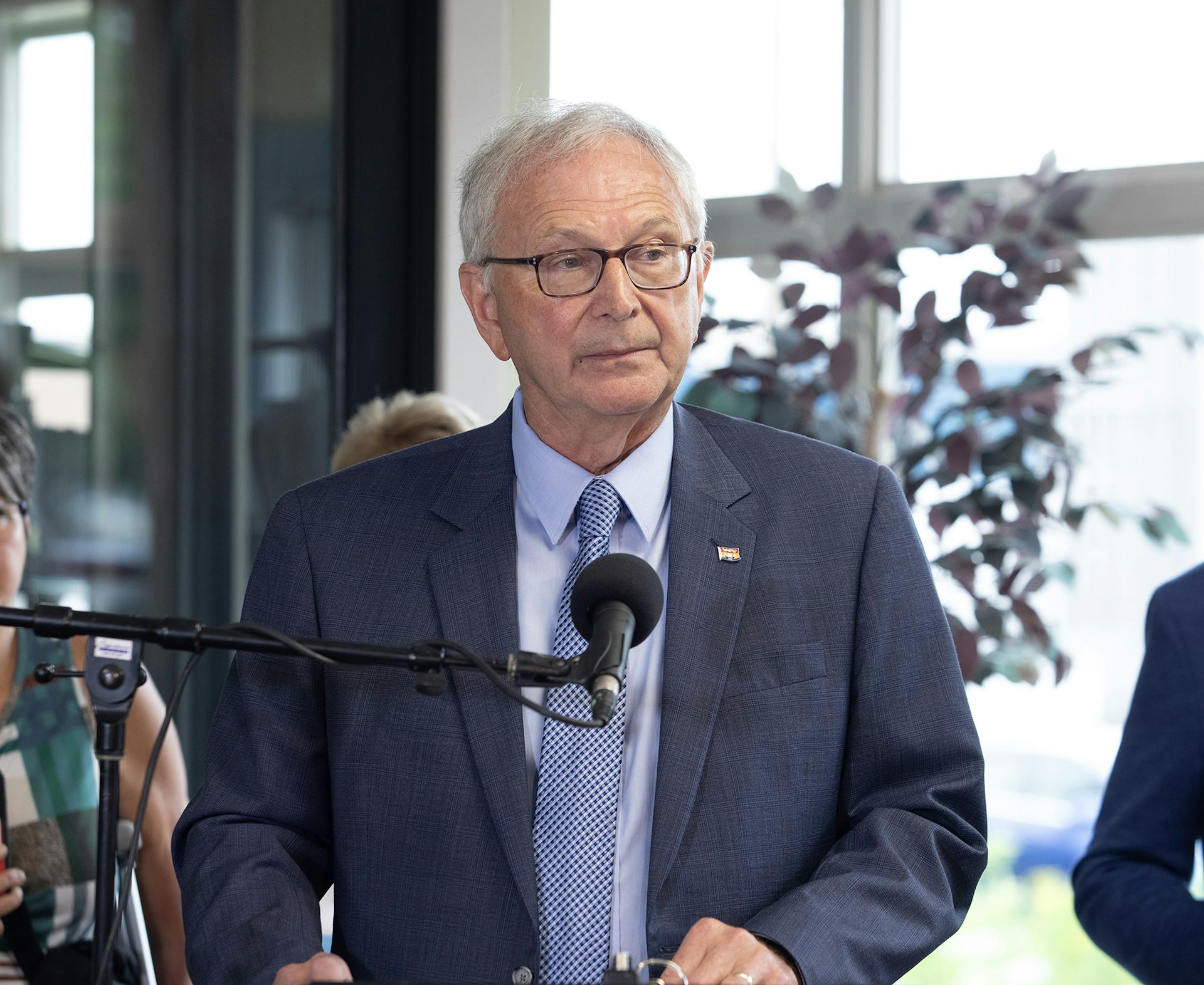 A man in a grey suit with grey hair and glasses stands behind a microphone.
