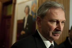 A man with a moustache and a painted portrait behind him.
