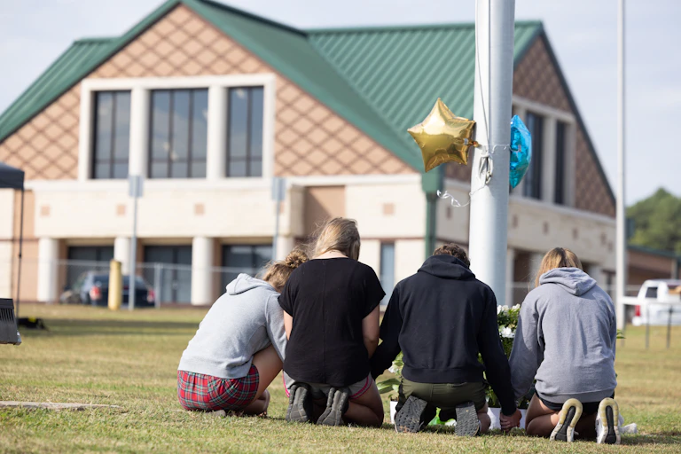 Students kneel at a makeshift memorial near a flagpole outside of a high school.