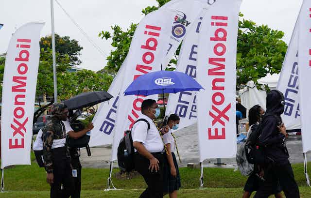 People with umbrellas and backpacks walking past ExxonMobil flags.