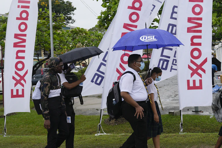 People with umbrellas and backpacks walking past ExxonMobil flags.