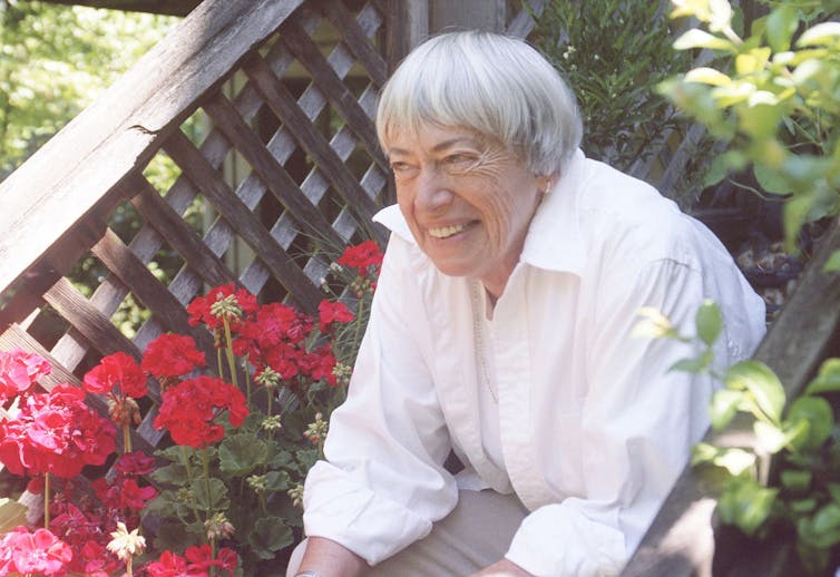 Smiling elderly woman with gray hair and a bowl haircut sits on steps surrounded by red flowers.