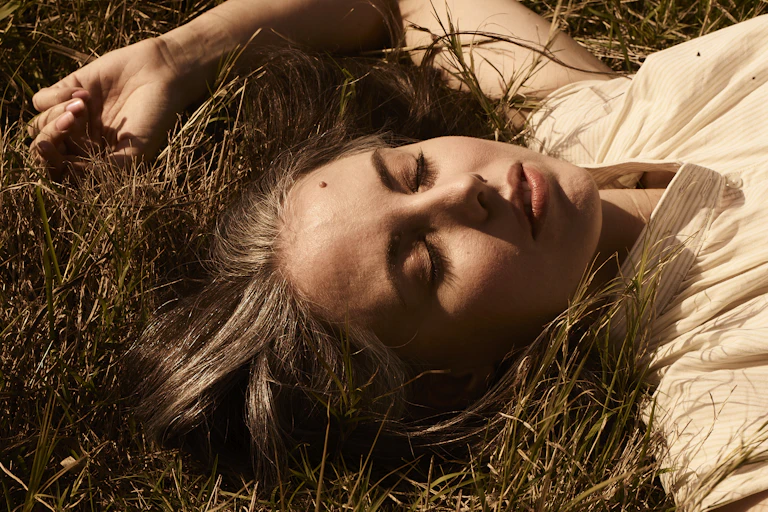 A women lies in dry grass with her eyes closed.