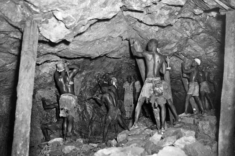 Black and white photo of a rocky tunnel propped up by poles, with shirtless men applying tools to the rockface