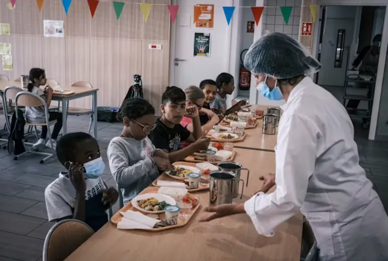enfants assis à une table dans une salle de cantine, une femme en blouse au premier plan