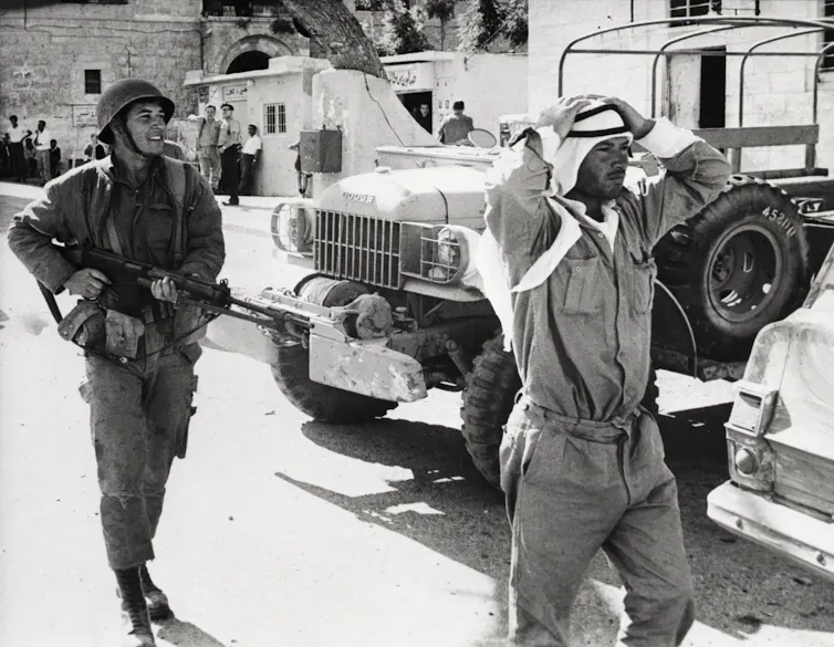 A man in Arab headscarf has his hands on his head while being followed by a man in army uniform carrying a gun.