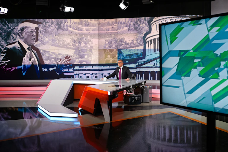 A man sits at a news desk surrounded by big screens.