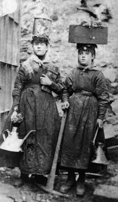 Dos mujeres con ropa de trabajo, cestos en la cabeza y aceiteras en las manos.