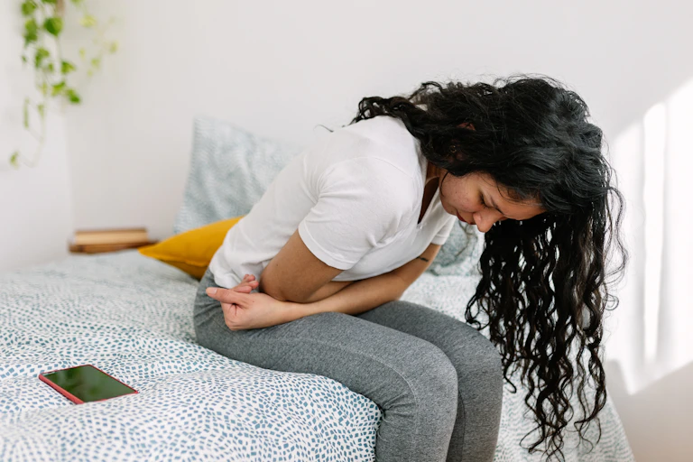 Young millennial woman feeling unwell and leaning over on a bed clutching her stomach.