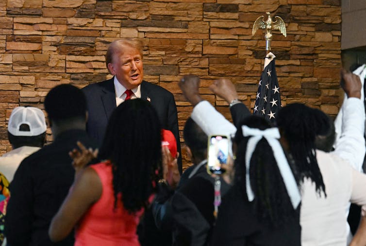 A middle-aged white man dressed in a business suit tries to talk with several Black people who have their fists raised in the air.