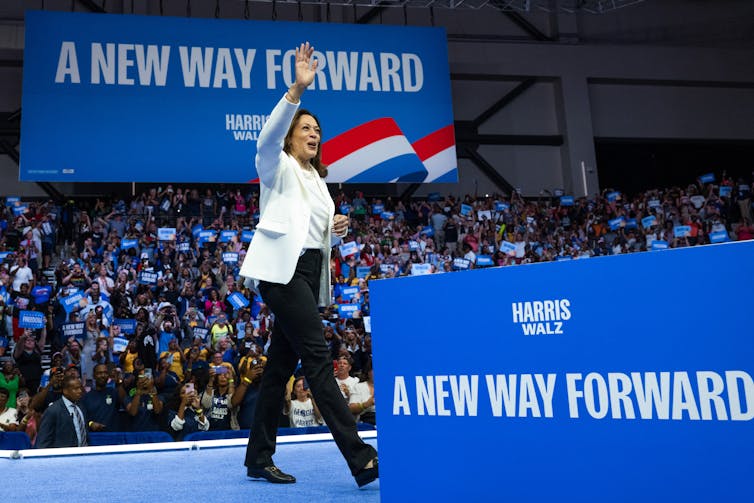 A woman wearing a white blazer and dark pants waves as she walks on a stage in front of a large crowd of people, with the signs 'A New Way Forward' around her in blue.