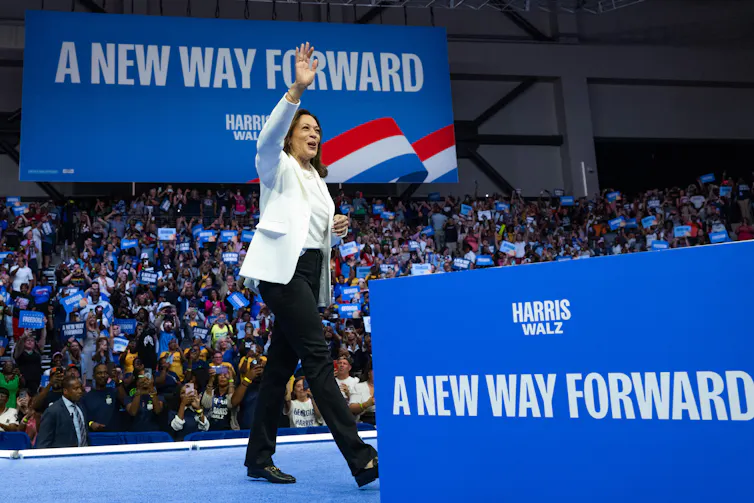 A woman wearing a white blazer and dark pants waves as she walks on a stage in front of a large crowd of people, with the signs 'A New Way Forward' around her in blue.