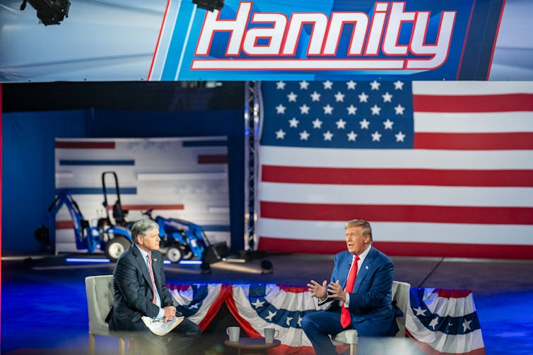 Two men face each other and are seated. There is a large American flag behind them and the word 'Hannity' on a screen.
