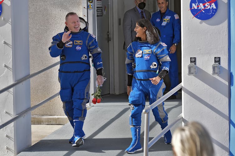 Astronauts Butch Wilmore and Suni Williams, wearing flight suits, walk down a ramp with the NASA logo in the background.