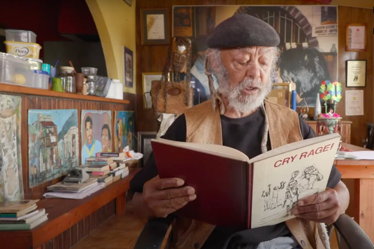 An elderly man in a beret with a great beard sits and reads from a book title 'Cry Rage'.