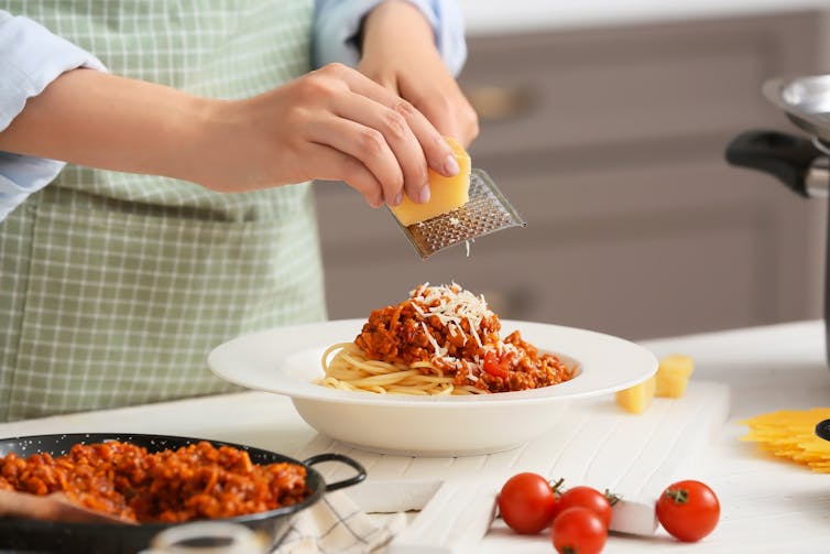 Woman's hands grating parmesan cheese into a bowl of spaghetti.