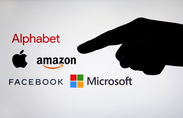Silhouette of a finger pointing at the logos of big tech companies.