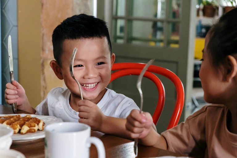 Toddler holding a knife and fork smiles at his sister
