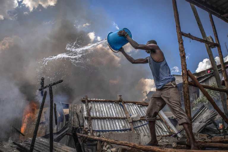 Man throws bucket of water towards flames and smoking metal structure