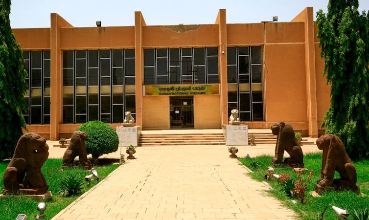 An ochre-coloured, flat building with ornate concrete window frames stands in a garden with ancient sculptures of lions lining the road up to it.
