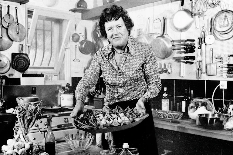 black and white photo of woman standing in cluttered kitchen holding a platter of food and smiling