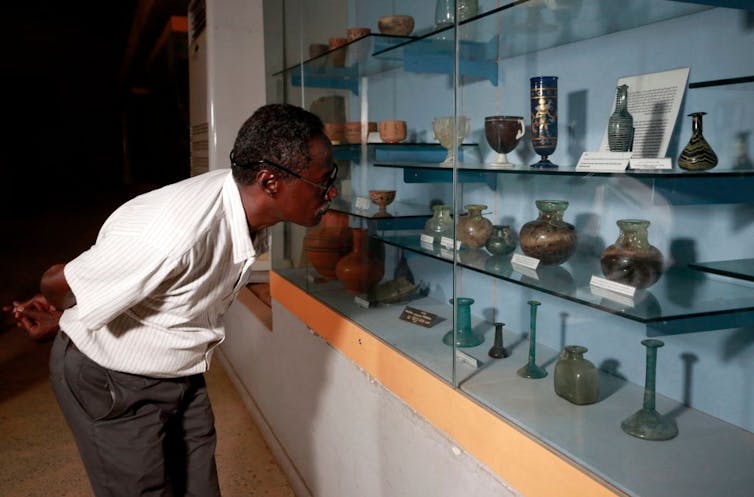 A man bends and peers into a glass cabinet containing numerous old pottery and ceramic objects.