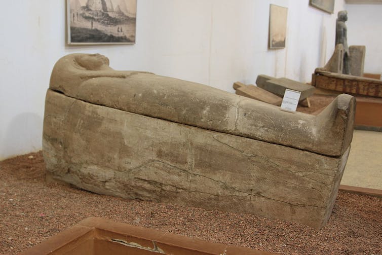 A tomb in the shape of a human that resembles a mummy rests on gravel, in a row of similar objects.