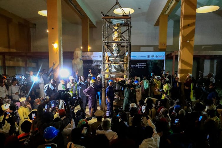 Workers are lit up by camera flashes as media throng in front of them where they stand with an enormous sculpture in a cage being moved by machinery.