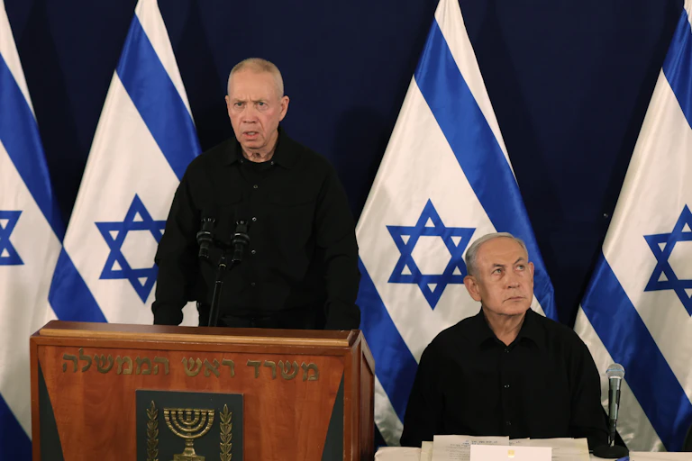 Yoav Gallant delivering a speech with Netanyahu sat beside him.
