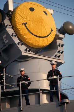 Two soldiers in uniform stand with their hands clasped behind their backs on the deck of a warship. A yellow smiley face looms over them.