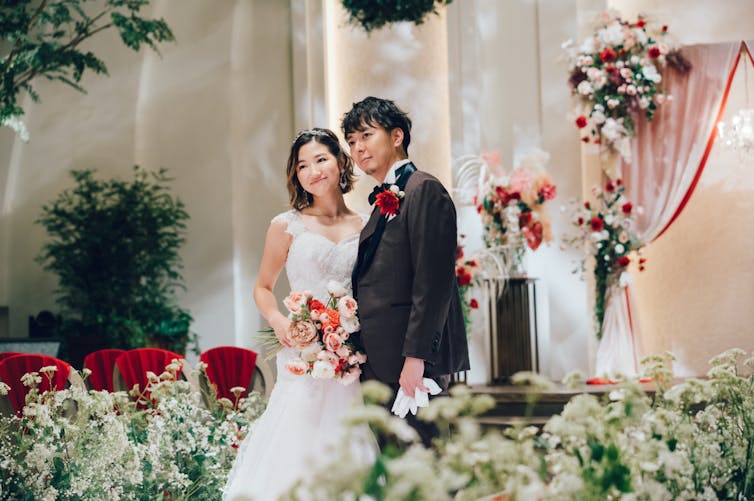 A woman in a white wedding dress holds a bouquet; a man in a black tuxedo stands close to her.