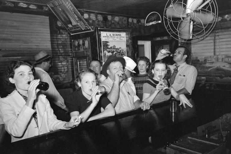 A handful of people stand in a tin-walled bar holding bottles up to their mouths in a black and white photo.