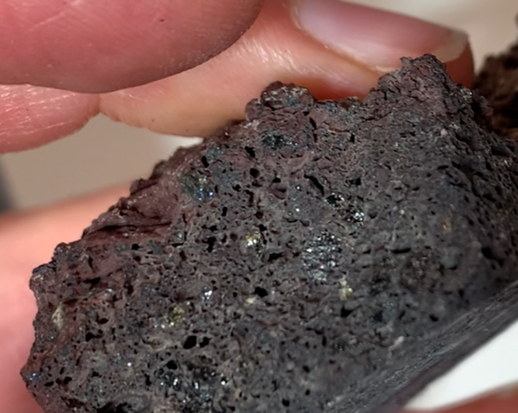Photo of a person's hand holding a lump of black volcanic rock with tiny sparkling crystals embedded in it.