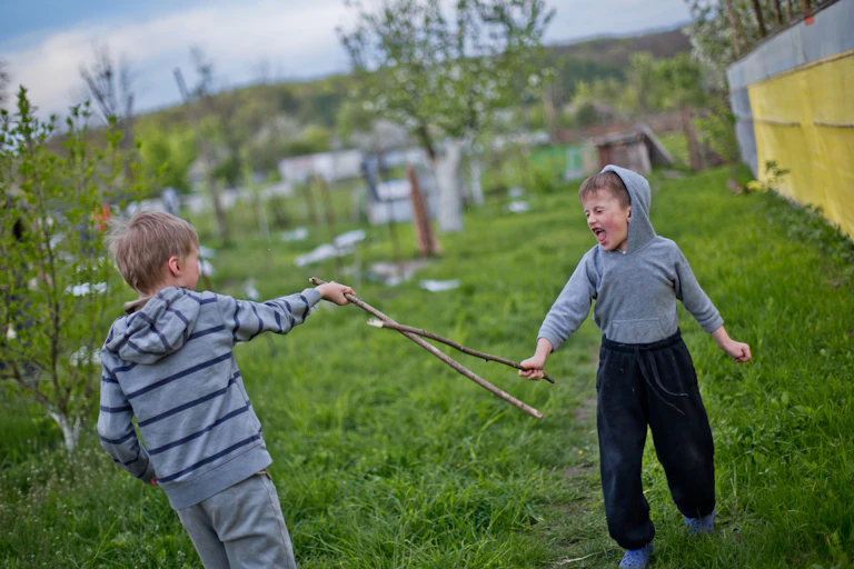 Two young boys fight with sticks on green grass. One boy shouts with his eyes closed.